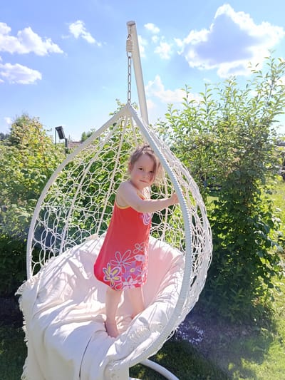 A young girl in a red dress with a floral pattern stands on a white blanket inside a white egg-shaped hanging chair in a garden. The chair is suspended by a white chain from a stand. The girl is looking at the camera with a slight smile. Behind her, lush green bushes and trees are visible under a blue sky with scattered white clouds. The ground is covered in grass. The image appears to be taken outdoors on a sunny day. This scene is consistent with a residential backyard in Shishkin Les, Russia.