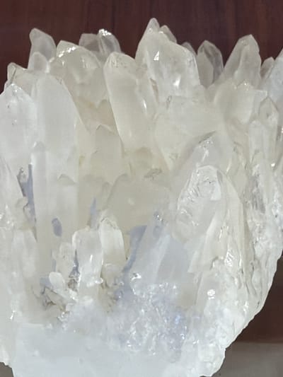 This is a close-up, medium-shot photograph of a cluster of clear, translucent crystals. The crystals are jagged and angular, with many facets reflecting light. They appear to be quartz or a similar mineral. The background is blurred and consists of a dark, grainy surface, possibly wood, on the left side, and a lighter, out-of-focus area on the right. There is no discernible activity or event taking place, as the image focuses solely on the mineral formation. The lighting is bright, highlighting the transparency and texture of the crystals. The image is devoid of any text or people. The location context provided is Akatovo, Russia, but there are no visual cues within the image itself to confirm this.