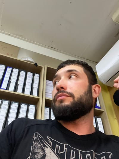 The image shows a close-up, low-angle, head-and-shoulders shot of a young adult male with dark hair, a beard, and brown eyes, looking to his right. He is wearing a black t-shirt with a graphic design. The background is an office environment with shelves filled with binders and folders. An air conditioning unit is visible on the wall above and to the right of the man's head. The lighting suggests an indoor setting with overhead lights. The image does not provide any information that would allow for the determination of the specific location within the city of Luzhitsy, Russia.