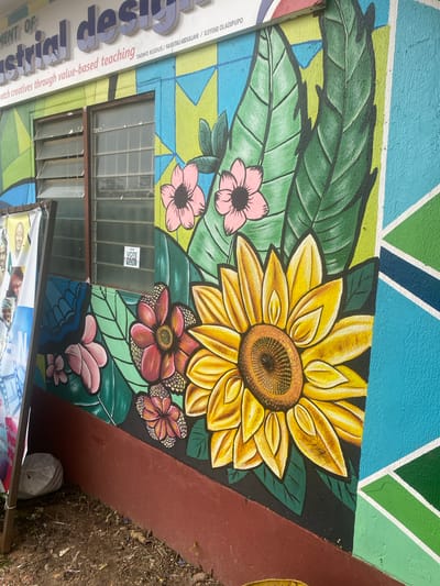 The image depicts the exterior wall of a building adorned with a vibrant mural in Asha, Nigeria. The mural features large, stylized flowers, including a prominent sunflower and several pink and red blossoms, interspersed with green leaves. The background is a dynamic geometric pattern of blue, green, and yellow. Above the mural, lettering on the building's facade reads "DEPARTMENT OF Industrial design" and "tch creatives through value-based teaching." A small sticker on a windowpane displays "VOTE PAPA 4 MARCH." To the left, a banner with portraits of men is partially visible, attached to a wooden support. The ground in front of the building is dirt with scattered debris.