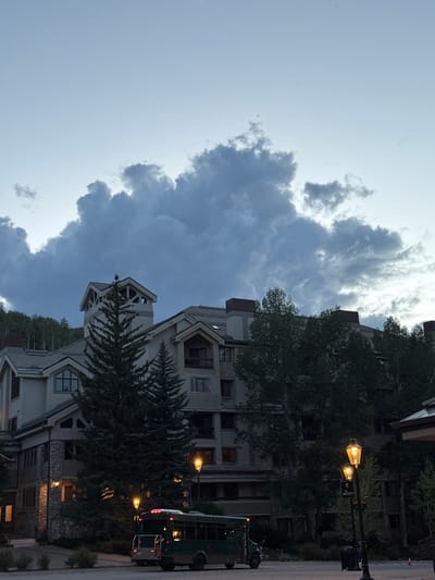 In Beaver Creek, Colorado, a large, multi-story building with a stone and stucco exterior is visible under a dramatic sky filled with dark, billowing clouds. The building has multiple windows and balconies, and a prominent tower with a lattice-like top extends upwards. Several evergreen trees are positioned in front of the building, with deciduous trees and a treeline visible further in the background. In the foreground, a dark green shuttle bus with "BEAVER CREEK" lettering on its side is parked on a paved area. Two ornate lampposts with illuminated globes stand near the bus. The scene suggests an evening setting, with the sky indicating approaching dusk or inclement weather.