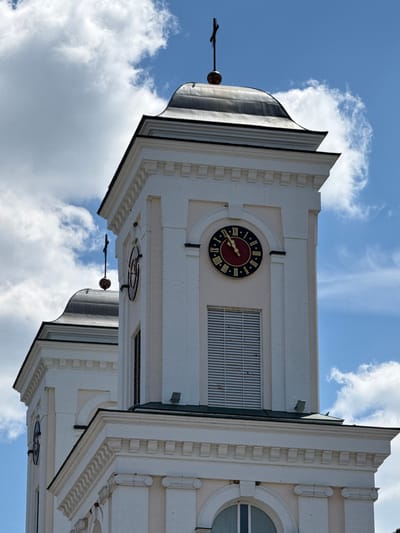 This is a vertical, eye-level shot of a white, multi-tiered building against a bright blue sky with scattered clouds. The building appears to be a clock tower, with at least two clock faces visible. The main clock face, in the center of the image, is dark red with Roman numerals and gold hands, indicating approximately 2:45. Another clock face is visible further down the tower, partially obscured. The architecture features decorative elements such as horizontal banding, rectangular patterns, and curved molding. A pointed, dark spire with a finial is visible on the left side of the frame, silhouetted against the sky. The overall impression is of a grand, old structure on a clear day. There is no visible text in the image.