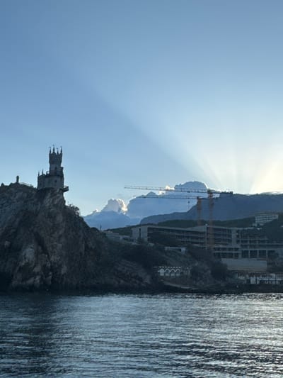 The image shows the Swallow's Nest castle perched on a cliff overlooking the sea, likely in the Гаспринский поселковый совет area of Ukraine. Construction cranes are visible in the background, suggesting development activity. The sky is clear with sun rays emanating from behind a cloud formation. The sea is calm with visible texture on the surface.