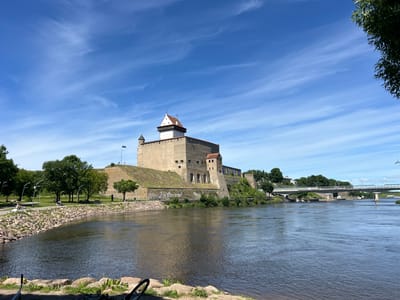 A wide, calm river flows in the foreground, reflecting the blue sky and wispy white clouds. On the far bank, a large, ancient stone castle with a white, pointed roof sits atop a grassy hill. The castle has several towers and defensive walls, and an Estonian flag flies from one of its turrets.
To the left of the castle, a park area with green grass and trees is visible. Several people are enjoying the sunny day, with some sitting on benches and others walking or cycling along a paved path. Lampposts line the path, and large rocks form a border between the park and the river.
Further in the distance, a modern bridge spans the river, connecting the two banks. Trees and buildings can be seen on the far side of the bridge. The overall scene suggests a warm, pleasant day in a historic European city, likely Narva, Estonia, given the castle's appearance and the presence of the Estonian flag. The presence of a bicycle in the foreground indicates it might be a popular spot for outdoor recreation. The sunlight and clear blue sky suggest it is daytime, likely late morning or early afternoon.