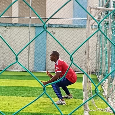 In Koko, Nigeria, an adult male is depicted on an artificial turf soccer field, viewed through a blurred green chain-link fence. The man has dark skin and short dark hair, and is positioned in a low crouch, leaning forward with open hands raised in front of him, suggesting a ready or defensive posture. He wears a red short-sleeved athletic shirt with a white Adidas logo and an "expedia" sponsor logo on the right chest, dark blue athletic pants with three light blue stripes down the sides, and light-colored athletic shoes. His head is turned to the left of the frame, and his mouth is slightly open. The field features alternating stripes of light and dark green artificial grass. In the background, a light-colored wall with vertical blue sections is visible, topped by a gray chain-link fence and additional building structures. A white metal soccer goal frame with a green net is partially visible on the far right.