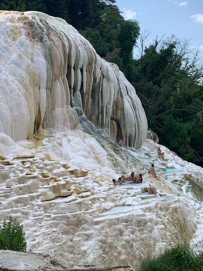 The image shows a group of people enjoying a natural hot spring at Bagni San Filippo, Italy. The setting is a unique geological formation characterized by white, mineral-rich deposits that have created tiered pools and cascading waterfalls. The water appears clear and inviting, with a light blue hue in some areas.

Several individuals are visible, some wading in the shallow pools, others sitting on the mineral formations, and one person appears to be in the process of diving or jumping into the water from a higher ledge. The people are dressed in swimwear, suggesting a recreational and leisurely activity.

The mineral deposits have a layered, textured appearance, with variations in color ranging from bright white to off-white and hints of earthy tones. The natural contours of the formations create an organic and sculpted landscape. Lush green trees and foliage are visible in the background, framing the hot springs and adding a sense of natural beauty to the scene. The sky above is a light blue with scattered white clouds, indicating fair weather, likely during the daytime. The overall mood conveyed is one of relaxation, fun, and enjoyment of a natural wonder.