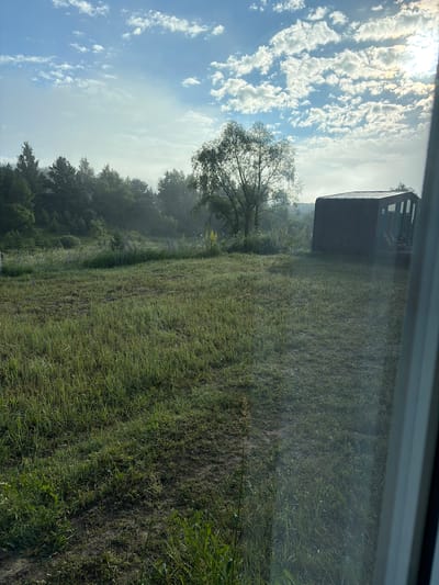 The image depicts a rural scene with a grassy field in the foreground, a line of trees in the midground, and a cloudy sky with the sun peeking through in the background. To the right of the midground, a dark, box-like structure with windows is visible. The environment appears to be a natural landscape with some signs of human presence. The specific location in Avchurino, Russia, is not discernible from the visual information provided. There are no people or identifiable actions in the image.