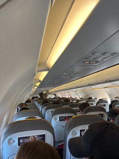 The image shows the interior of an airplane cabin, looking forward from the back of the cabin, from the perspective of a passenger seated in the aisle seat. There are rows of passengers seated in front of the viewer, all facing forward. The seats are grey with yellow accents. The aisle is visible on the viewer's left, with a passenger seated in the aisle seat in the row in front of the viewer. Several passengers are visible in the image, including one wearing a black baseball cap. The overhead lights are turned on. There is an advertisement on the back of the seat in front of the viewer, featuring a woman wearing a red top. The overhead bins are visible. The location is unknown.