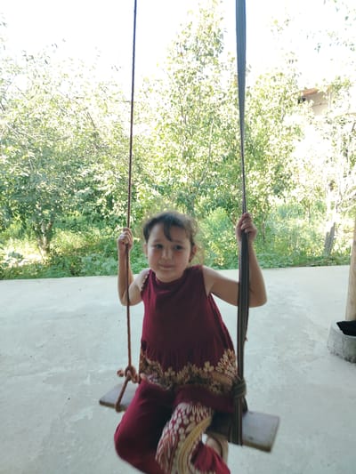 A young girl with dark hair and a red dress is sitting on a swing outdoors. She is holding onto the ropes of the swing with her hands. The swing set is made of wood and rope. The background is filled with green trees and foliage, suggesting a natural or rural setting. The ground appears to be paved or concreted. The sunlight is bright, indicating a daytime scene. The girl is smiling and looking towards the camera. The image was taken in Shovot, Uzbekistan.