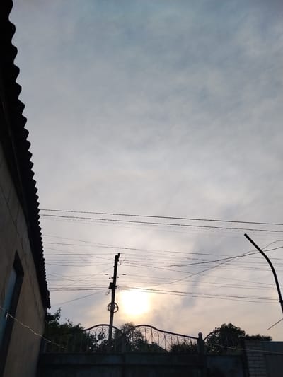 The image depicts an outdoor scene, likely in Vulcănești, Moldova, with a cloudy sky suggesting overcast or dawn/dusk conditions. The sun is partially visible, peeking through the clouds, creating a soft, diffused light. In the foreground, a portion of a building with a corrugated roof is visible on the left. A dark fence with decorative metalwork runs horizontally across the lower part of the frame, separating the foreground from the background. Behind the fence, lush green trees are silhouetted against the sky. Several power lines crisscross the scene, strung between utility poles, one of which is prominent in the mid-ground. A dark, curved pole, possibly a street light, is visible on the right side. The overall mood is tranquil and somewhat muted, due to the cloudy sky and diffused sunlight. No people are visible in the image. There is no visible text.