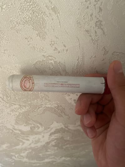 A hand holds a white cigar with a red tip against a textured, light-colored wall. The cigar has a circular pink and red label on one end and text printed on its body. The textured wall has a swirling, abstract pattern. The scene is indoors, likely in a domestic setting. There is no information available to determine the location within the city of Istaravshan, Tajikistan.