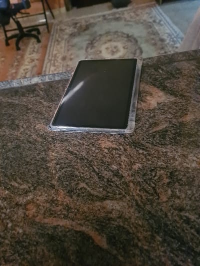 A tablet rests on a marble surface with a patterned texture. The screen is dark, suggesting it's off or displaying a black background. A corner of a patterned rug is visible in the background, along with parts of a chair. The photo is taken from a slightly elevated angle, tilted to the left.