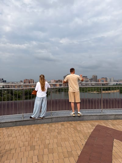 Two individuals, a woman and a man, stand on a paved elevated walkway overlooking a cityscape. The woman, seen from behind, has long blonde hair and wears a white t-shirt and wide-leg light blue jeans, carrying a brown handbag. The man, also viewed from behind, wears a light beige t-shirt and matching shorts, with his right arm raised as if gesturing. Both are leaning on a metal railing that separates the walkway from the view. Beyond the railing, a river or body of water is visible, with a tree-lined bank and a cluster of modern buildings forming the skyline. The sky is overcast with gray clouds. The walkway is paved with light brown and reddish-brown square tiles. This scene is located in Tarlashi, Russia.