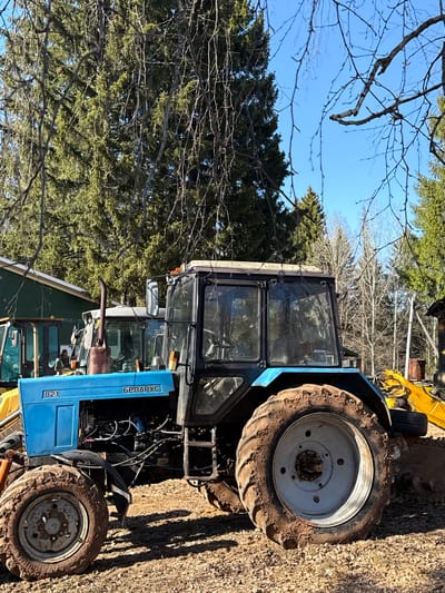 The image shows two blue tractors in a rural environment, likely in Галево, Russia, given the Cyrillic script on one of the tractors ("БЕЛАРУС"). Both tractors appear to be Belarus 82.1 models, with one partially obscured by the other. The tractors have dirty tires, suggesting recent use in muddy conditions. One tractor has a full cab, while the other's engine is exposed. There are green trees and bare branches in the background, indicating a possible forest or wooded area. Part of a yellow construction vehicle can also be seen. The ground is covered with dry leaves and dirt. The sky is blue and clear.