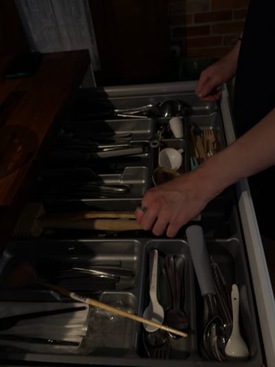 The media file shows a close-up, overhead view of an open kitchen drawer filled with various kitchen utensils. A person's left arm and hand are visible, reaching into the drawer. The hand is adorned with a silver ring. The drawer contains a cutlery organizer with compartments holding spoons, forks, knives, and other kitchen tools. Some items appear to be made of stainless steel, while others are wooden or ceramic. A small wooden mallet or tenderizer is also present. The scene is dimly lit, suggesting an indoor setting, possibly a kitchen. The background includes a brick wall and a dark wooden drawer front. The overall impression is one of domestic activity, with someone organizing or selecting utensils. The time of day and weather are not discernible from the image. There is no visible text in the media file. The location context provided is Słupsk, Poland, but this information is not directly evident from the image itself.