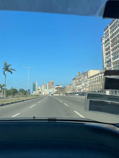 The image presents a sun-drenched urban street scene in Luanda, Angola, captured from inside a vehicle looking forward and slightly to the left. The sky is a vast, cloudless blue, indicating bright daylight, likely midday or early afternoon.
On the left side of the frame, a continuous stretch of multi-story buildings lines the street, receding into the distance. The closest structure on the far left is a very tall, modern building with numerous windows or balconies. Further along, other buildings, some appearing to be under construction or featuring extensive external structures, are visible. A small, colorful flag with red, green, and black stripes can be seen on one of these more distant buildings, possibly the Angolan flag. The word "BANK" is clearly visible on a building further down the street, and letters resembling "ANB" or "AMC" are discernible on another.
The right side of the image is dominated by a wide, paved road with multiple lanes delineated by solid and dashed white lines. The road extends straight into the horizon. A median strip, featuring sparse green vegetation and streetlights, separates the road from the buildings or perhaps a parallel carriageway. Several palm trees are scattered along this median and further down the roadside, contributing to a tropical aesthetic.
In the immediate foreground on the far right, a dark gray or black section of the vehicle's interior, likely a door panel or dashboard, partially frames the scene. A small, rectangular white object, possibly a sticker or card, is attached to this interior part, bearing faint, illegible text. The overall scene depicts a modern cityscape under clear and sunny weather conditions.