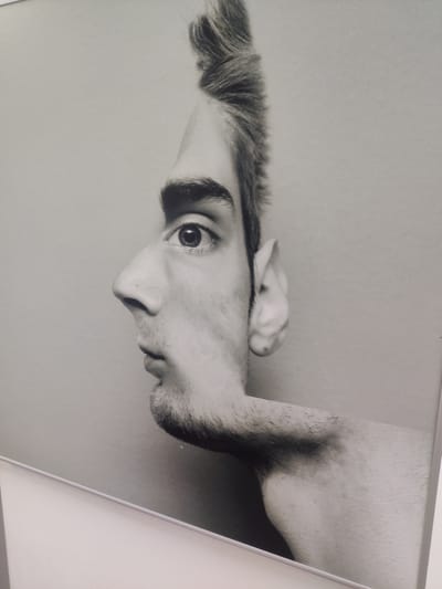 The image displays a black and white photographic artwork that utilizes optical illusion to depict two faces in profile. One face is viewed from the left, showing an eye, nose, mouth, and beard stubble, with spiky hair extending upwards. This face appears to be part of a larger figure. The second face is viewed from the right, with its profile formed by the negative space around the first face, revealing an ear. The artwork is framed and appears to be mounted on a light-colored wall. The background is a plain, neutral tone. The lighting is soft and even, characteristic of an indoor exhibition space. The artwork is positioned in a corner, with the bottom edge of the frame and a white surface visible in the foreground. There is no information within the image that indicates its location in Thessaloniki, Greece.