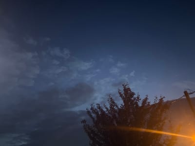 The image captures a nighttime scene with a deep blue sky above a dark, silhouetted tree in the foreground. Wispy, lighter clouds are scattered across the sky, illuminated by an unseen light source. A utility pole with wires is visible on the right side of the frame, and a bright orange light flares across the lower right corner of the image. The overall impression is one of twilight or early night in an outdoor, possibly suburban or rural, setting. No people or specific actions are depicted. The location is identified as Dubovoye, Russia.