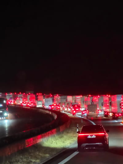 The image depicts a nighttime traffic jam on a highway, likely the A3, near Uettingen, Germany. A dark-colored SUV with illuminated taillights is in the foreground, facing away from the viewer and traveling in the right lane. Behind the SUV, numerous trucks are lined up, their red taillights creating a vibrant glow against the dark asphalt. The highway appears to be multi-lane, with vehicles moving slowly or stopped. The environment is characterized by the darkness of night, with the only significant light sources being the vehicle lights and some roadside illumination. The scene conveys a sense of congestion and standstill traffic.