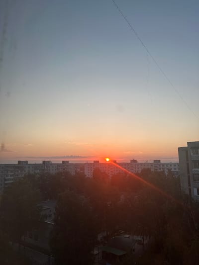 A wide-angle, eye-level shot captures a sunrise over a residential area. The sky transitions from a pale blue at the top to a soft orange and pink gradient near the horizon. A thin, dark wire with a wavy pattern stretches diagonally across the upper portion of the sky. In the middle ground, a line of multi-story apartment buildings, characteristic of Soviet-era architecture, stretches across the frame. The buildings are light gray with numerous windows. A low-lying layer of fog or haze obscures the ground beyond the buildings. In the foreground, dense green trees partially obscure the lower levels of the buildings. The sun, a bright orange orb, is visible just above the horizon, casting a warm glow and a lens flare across the scene. The overall impression is a tranquil urban landscape at dawn. The specific location is the city of Bakhchisarai, Russia.