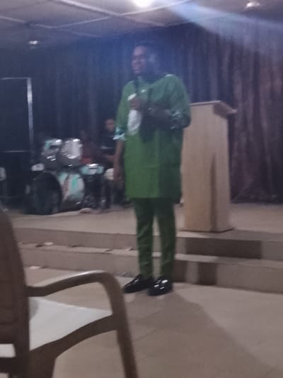 A man stands on a raised stage, addressing an unseen audience, holding a white object, possibly a microphone. He wears a green tunic and matching trousers with black shoes. A wooden podium stands to his right. A drum set is visible to the left of the man. A chair is in the foreground and brown curtains serve as a backdrop. The lighting is dim.