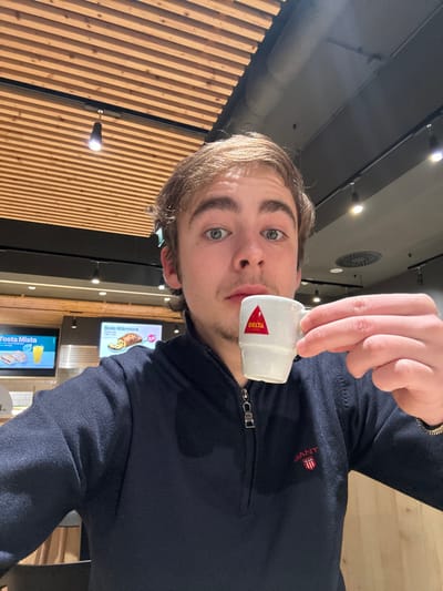 A fair-skinned young man with light brown hair and blue eyes holds a small, white, handled espresso cup with a red triangular logo. He is wearing a dark navy blue half-zip sweater with the name "GANT" embroidered on the right side. The background suggests he is indoors, possibly in a cafe or restaurant, with wooden slats on one wall, ceiling lights, and menu boards visible. The image is tilted, giving a slightly disorienting perspective.