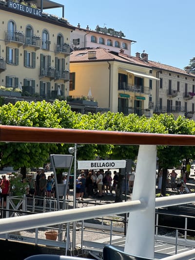 The image shows a waterfront scene in Bellagio, Italy, with several buildings in the background, including the "HOTEL DU LAC" and "HOTEL BELLAGIO". In the foreground, a group of people are gathered near a sign that reads "BELLAGIO". There are trees and a railing in the mid-ground. The scene appears to be a public area, possibly a ferry terminal or a promenade. The overall atmosphere is bright and sunny, suggesting a pleasant day.