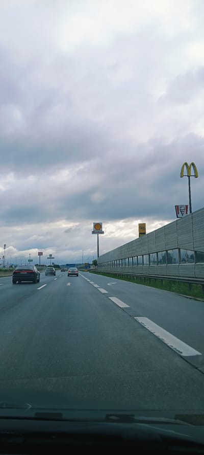The image depicts a highway scene from the perspective of a driver. The road is paved and has multiple lanes marked with white lines. Several cars are driving on the highway. On the right side of the road, there is a sound barrier made of grey panels with windows. Above the sound barrier, there are several billboards. One billboard displays the McDonald's logo, and another shows a KFC bucket with Colonel Sanders' face. Further in the distance, a Shell gas station sign is visible. The sky is overcast with grey clouds. The image appears to be taken in the city of Ansfelden, Austria, as indicated by signs for "Ansfelden" on the Shell billboard and a sign with the text "Raststation" which translates to "rest stop" or "service area" in German, commonly found at highway rest stops in Austria.