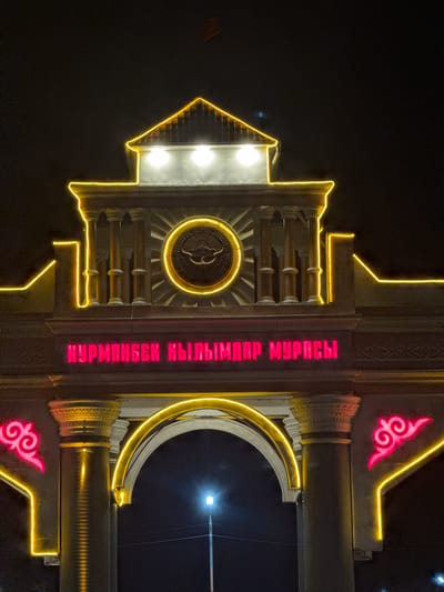 The image shows an illuminated archway at night in Suzak, Kyrgyzstan. The archway is adorned with yellow neon lights that outline its structure and a prominent crest with a sun and mountains in the center. Above the arch, red neon letters spell out a message in Cyrillic script. The background is dark, indicating nighttime, with a street light visible in the distance. The architecture appears to be traditional or ceremonial. There are no people or animals visible in the frame.