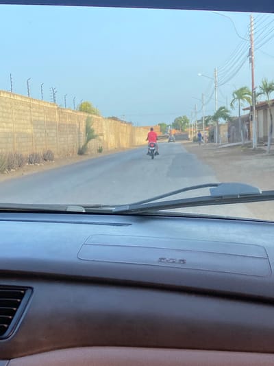 The image depicts a daytime scene from inside a vehicle, looking out onto a road in La Cruz del Pastel, Venezuela. The sky is bright and clear.

In the foreground, a person wearing a red shirt is riding a motorcycle away from the viewer on what appears to be an asphalt or paved road. Further down the road, two other individuals are visible in the distance, one of whom is wearing blue.

The left side of the road is bordered by a long, concrete block wall, behind which some sparse vegetation and palm trees are present. Utility poles with numerous overhead wires line the side of the road, stretching into the distance.

The interior of the vehicle is visible on the right, showing a dashboard, part of a windshield wiper, and an air vent. Faintly embossed text on the dashboard reads "SRS AIRBAG". The overall atmosphere suggests clear, sunny weather in a semi-urban or rural setting.