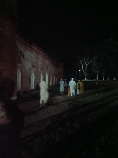 The image depicts an outdoor nighttime scene with a long, brick building on the left and a group of people walking away from the viewer on a path. The building, likely part of the historic Khan Jahan Ali's Tomb complex in Bagerhat, Bangladesh, is constructed of red brick and features a series of arched openings. The individuals are dressed in traditional South Asian attire, with at least two men wearing light-colored garments, and one person draped in a yellowish-brown shawl. The surrounding area appears to be a park or garden, with trees visible in the background and shadows cast across the ground, suggesting artificial lighting. The overall atmosphere is dark, with limited visibility, and the image quality is somewhat blurred.