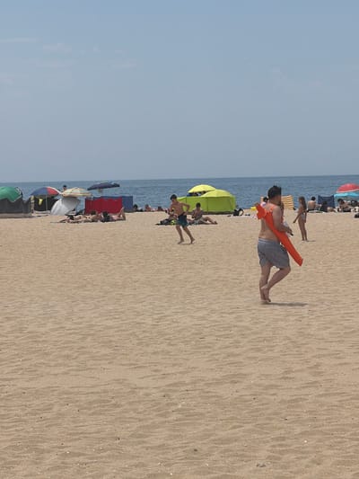 The image shows a crowded beach scene on a sunny day. In the foreground, a man in blue and white striped shorts is walking with an orange float slung over his shoulder. He is facing away from the camera. Further back, several people are sunbathing or relaxing under beach umbrellas and tents. Some individuals are visible in the ocean. The beach is sandy, with the ocean in the background under a clear blue sky. This scene is typical of Espinho's Praia da Baía, known for its lively atmosphere during summer months.
