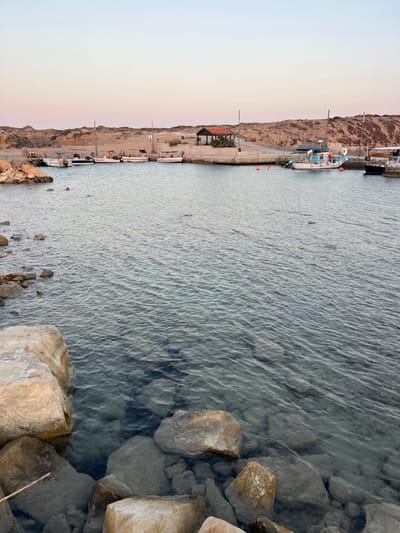 The image depicts a coastal scene at a harbor, likely in Akrotiri, as suggested by the location context. Several boats are docked along a concrete pier, with some appearing to be fishing vessels. The water is clear and calm, revealing rocks submerged beneath the surface near the shore. In the background, a rocky, arid hillside rises, characteristic of a Mediterranean landscape. The sky exhibits a gradient of pale blue and soft orange, indicating either dawn or dusk. The overall atmosphere is serene and quiet, with no people actively engaged in any visible activity. The time of day appears to be either early morning or late evening, due to the soft lighting and pastel colors in the sky. The clarity of the water and the rocky terrain provide specific geographical cues. One of the boats has visible text that reads "STAVROS".