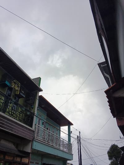 The image depicts an upward-angled view of an urban environment in Pinnwe, Myanmar, under an overcast, cloudy sky. On the left, a two-story structure is visible; the upper level features a light green facade with a balcony enclosed by a black, geometrically patterned metal railing. Yellow fabric items are draped over this railing. Below, a brown horizontal striped section is visible, with a partially legible sign at the extreme bottom left. Adjacent to this, a light teal-colored building presents a second-story balcony with a silver-colored, ornate metal railing and multiple windows. Its roofline features a dark brown soffit. From the right, a darker building's roof eaves and upper wall are partially visible, framing the sky. Numerous black electrical and communication lines crisscross the frame at various angles. A dark utility pole, equipped with multiple cross-arms and a dense cluster of wires, stands in the lower-middle right. A small amount of dark green foliage is present at the bottom right corner. No individuals are depicted in the scene.