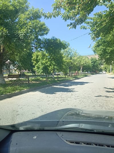 The image shows a street in Izberbash, Russia, on a sunny day. The street is lined with trees and a fence. There are a few cars on the street and a building in the background. The perspective is from inside a car, looking out. The dashboard and windshield of the car are visible in the foreground.