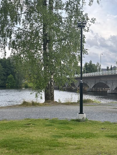 The image captures a scene in Vammala, Finland, with a focus on a birch tree in the foreground, its slender white trunk and green foliage dominating the left side. To the right of the tree, a black lamppost with two lights stands tall. Beyond the tree and lamppost, a wide river flows, its surface rippled by a gentle breeze. In the background, a stone bridge with multiple arches spans the river, its structure appearing old and well-maintained. A white railing lines the top of the bridge, and beyond it, various buildings and trees are visible. The sky is overcast, with clouds suggesting a partly cloudy day. The foreground consists of a gravel path adjacent to the river, followed by a lush green lawn. No people are visible in the scene.