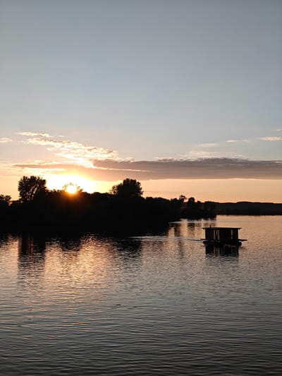 This image captures a sunset or sunrise over the Kama River in the city of Nikolo-Beryozovka, Russia. The scene is dominated by a body of water reflecting the ambient light and a warm sky.
The upper portion of the frame features a sky transitioning from a light, hazy blue-grey at the zenith to warm orange and pink hues near the horizon. Several diffused, elongated clouds are visible, notably above the radiant sun and in the mid-upper right quadrant. Small, dark specks, possibly distant birds or insects, are faintly discernible in the sky.
The sun is positioned on the left side of the horizon, partially obscured by a dense line of silhouetted trees. Its bright, disc-like form emits a strong, localized glow, creating a sunburst effect through and above the foliage. Further to the right, a distant, gently undulating landmass or range of hills appears as a dark silhouette against the sky.
The lower two-thirds of the image consist of the Kama River, its surface covered in small, consistent ripples that create a textured pattern. The water reflects the warm colors of the sky and the sun's glow, particularly near the horizon line, resulting in streaks of orange and yellow. The dark reflections of the silhouetted trees are also visible on the water's surface.
In the mid-ground on the right side of the river, a dark, rectangular floating structure is present. This appears to be a small, rustic house or shack built upon a raft or pontoons. It exhibits visible roof and wall elements. A subtle disturbance or wake trails behind the structure, indicating it is either in motion or has recently been moving, though no propulsion mechanism is explicitly visible. No individuals or clearly identifiable animals are present in the frame.