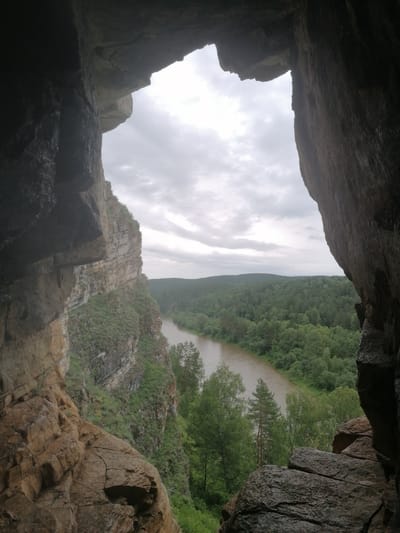 This media file depicts a view from inside a cave or rock overhang, looking out at a scenic natural landscape. The foreground is dominated by the rough, textured rock formations that form the mouth of the cave. These rocks are a mix of browns and grays, with visible cracks and variations in their surface.
Through the opening, a wide, muddy river flows through a valley. The river is bordered by lush, green forests that cover rolling hills. The trees are dense and appear to be a mix of deciduous and coniferous types. The sky is overcast with gray clouds, suggesting a cloudy or possibly rainy day. The overall lighting is subdued.
The scene appears to be a natural, undeveloped location, likely a natural park or wilderness area. The perspective suggests the viewer is standing within a geological formation, possibly a cave or a natural arch, observing the expansive view. There is no text visible in the image. The mood conveyed is one of natural grandeur and perhaps a sense of awe or peace inspired by the vastness of the landscape. The time of day appears to be daytime, but the overcast sky makes it difficult to determine a precise hour. The location is identified as Lagerevo, Russia.