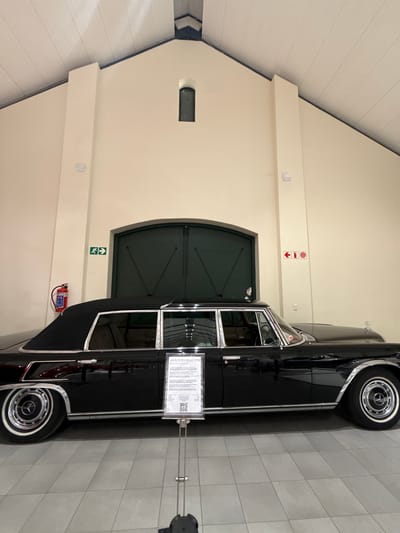 The image shows a black Mercedes-Benz limousine, likely a 1970s model, parked inside a building. The building appears to be a museum or exhibition hall with cream-colored walls, a high ceiling, and light gray tiled floors. The location could be in the Stellenbosch Local Municipality, South Africa. A pair of large, dark green doors is visible in the background. There are signs on the wall indicating a fire safety system, as well as an accessibility sign. A fire extinguisher is mounted on the wall near the car. A sign stands next to the vehicle that describes the car and has a QR code. The car itself appears to be well-maintained and is likely part of a collection or exhibit. There are no people visible in the image.