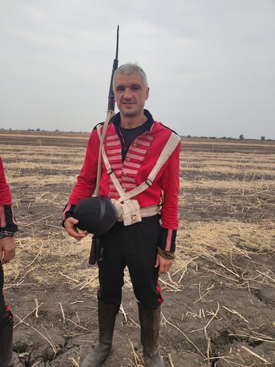 The image displays an outdoor scene in a dry, harvested agricultural field located in Kottalapalli, India, under an overcast sky. A Caucasian adult male, appearing approximately 30-45 years old with short grey hair and light facial stubble, stands predominantly in the center, looking directly at the camera with a slight smile. He is attired in a historical military-style uniform featuring a red long-sleeved jacket with horizontal black and light pink/red stripes on the chest and cuffs, black trousers, and dark brown knee-high boots. A white cross belt extends over his left shoulder, supporting a light-colored canvas pouch positioned on his right hip. A rifle, fitted with a bayonet, is slung over his left shoulder, its barrel extending upwards. The male holds a black shako-style hat in his right hand. A partial view of a second individual, wearing a similar red sleeve, black trousers, and dark boots, is visible on the far left of the frame. The ground consists of dry, cracked, dark brown soil mixed with scattered light-colored straw and crop stubble. The background features a flat, expansive field with a distant line of sparse trees against the grey sky.
