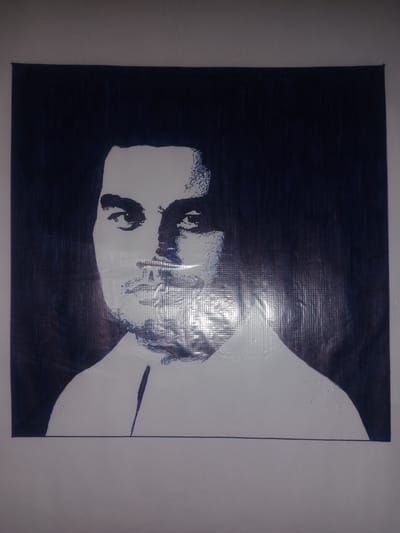 The image is a black and white portrait of a man. The portrait is on a dark background and appears to be a drawing or painting. The man is looking directly at the viewer. The portrait is mounted on a wall. The location is identified as Gulbag, Uzbekistan.