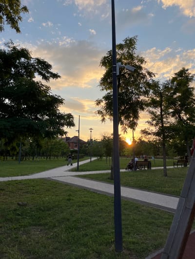 This vertical, eye-level, full shot captures a sunset scene in a park, likely within Taganrog City District, Russia. The sky is a blend of blue and warm hues of orange and yellow, with scattered clouds. In the foreground, a dark gray lamppost stands vertically on the right side of the image, equipped with a security camera. A paved pathway, a lighter gray with darker borders, winds through the park, splitting into two directions. To the left, a person on rollerblades is visible, moving away from the camera. The park features lush green grass and several trees with dark green foliage. In the middle ground, a house with a reddish-brown roof is visible behind the trees. Further back, on a bench, two adults are seated, with a stroller beside them. Another bench is visible further to the right. The overall ambiance is serene and indicative of evening leisure activities in a park setting.