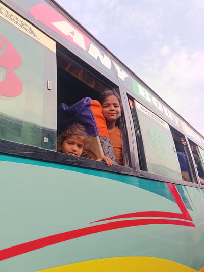 This image depicts a light teal and yellow passenger bus in Kottalapalli, India, against an overcast sky. Two young females are prominently visible looking out of an open window section on the left side of the bus.
The bus features include a light teal body with a yellow lower section, separated by a red striped design. Black window frames surround multiple windows. Visible text on the upper bus body includes "KANY" in gold letters on the left, a large pink stylized letter resembling an 'A' or an arrow, and "BODY" in green letters further right. Above one window, the word "DISCOTHEQUE" is printed. A small, silver rectangular vent is present on the bus body, to the left of the main window where the subjects are. A sticker with "SM K" in a white box is adhered to the lower left portion of the window glass.
The primary subject on the right is an older girl, estimated to be 7-10 years old, with light brown skin and dark hair. She wears an orange top and has a visible patterned garment below it. She is smiling, looking directly forward, with her left hand resting on the window sill.
To her left, partially obscured by a dark blue/purple fabric (possibly a shawl or garment), is a younger girl, estimated to be 3-5 years old. She has curly dark hair, a pink hair clip on her left side, and light brown skin. Her head and shoulders are visible as she looks forward with a neutral expression.
Further to the right, through another bus window, a person's arm and part of a blue garment are visible, suggesting other passengers. The sky in the background is uniformly bright and light grey, indicating an overcast daytime.