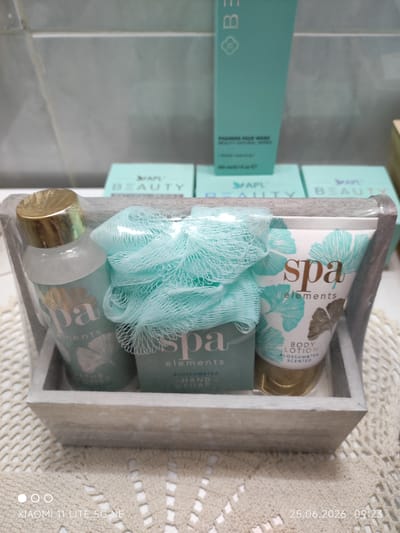 This media file displays a close-up shot of a spa gift set placed in a light-colored wooden basket. The set includes a small bottle with a golden cap, a blue mesh bath pouf, a bar of soap, and a tube of body lotion. The packaging of all items within the set features a soft blue and green color palette with delicate leaf-like patterns. The text "spa elements" is prominently displayed on the soap and body lotion, with additional text indicating "BODY LOTION BLOSSOM&TEA SCENTED" on the lotion tube and "HAND SOAP BLOSSOM&TEA" on the soap. In the background, two teal boxes with the text "APL BEAUTY" are visible, suggesting the brand of the products. The setting appears to be indoors, possibly a bathroom or a retail display, with a tiled surface visible in the upper portion of the image. The lighting suggests it might be daytime. The overall presentation evokes a sense of relaxation and self-care. A timestamp in the lower right corner indicates "25.06.2026 09:23". The bottom left corner shows "XIAOMI 11 LITE 5G NE", identifying the device used to capture the image.