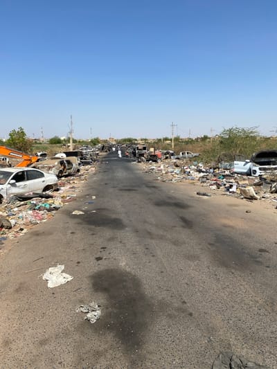 The image shows a street in Tayba Alhasanab, Sudan, located at latitude 15.36 and longitude 32.528. The street is littered with debris, including burned-out vehicles and scattered trash. There are two people in the distance, walking away from the camera. The street is paved and runs straight ahead, with a grassy median separating the two sides. The sky is clear and blue, with no clouds visible. There are power lines visible overhead. The scene is indicative of destruction and abandonment.