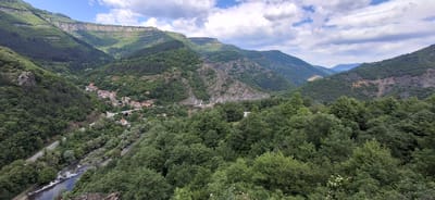 The scene depicts a lush mountainous valley in Bov, Bulgaria. A small village with red-roofed buildings is nestled within the valley alongside a river and a road. A bridge spans the river near the village. The surrounding mountains are covered in dense green vegetation under a cloudy sky.