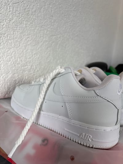 The media file contains a close-up, rotated shot of a white Nike Air Force 1 sneaker. The sneaker is made of white leather with white laces. The iconic Nike Swoosh logo is visible on the side of the shoe, and the word "AIR" is embossed on the midsole. The background is a textured white surface, possibly a wall or a backdrop. A portion of a clear plastic bag with red accents is also visible on the right side of the image. The lighting appears to be artificial, and the overall scene is neutral and clean, focusing on the product. No people are visible in the image, and the activity depicted is the presentation of the sneaker. No time of day, weather, or specific emotional cues are discernible. There is text on the shoe, including "AIR" and a logo that appears to be the Nike Swoosh. There might be some faint text on a label or tag at the top of the shoe, but it is not clearly legible.
