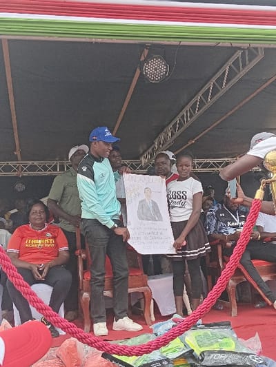 A group of people are gathered on a stage. One person holds a sign with a portrait and text, seemingly related to a political figure. The stage is constructed with metal framework, and there's decorative red and white rope along the front. Additional merchandise, possibly related to the event or the political figure, is stacked near the stage's edge. The people are dressed in casual clothing.