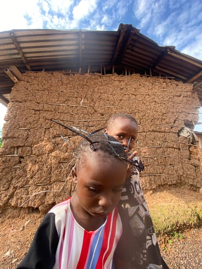 The image shows two young children, likely siblings, standing in front of a mud-brick wall under a partly cloudy sky. The wall, typical of rural architecture in places like Ufu Esioni, Nigeria, is constructed from brown mud mixed with straw or other fibrous material, reinforced with vertical wooden stakes. The roofline of a building, made of corrugated metal sheeting supported by wooden beams, is visible in the upper left of the frame.
The child in the foreground, wearing a striped shirt with red, white, and pink bands and a black lower section, is holding a pair of dark-rimmed glasses. The child's face is tilted downwards, and their expression is somewhat obscured. The second child, partially visible behind the first, is peeking over the first child's shoulder, also looking towards the camera.
The scene suggests a casual, everyday moment. The lighting indicates it is daytime, likely midday or afternoon, given the bright sunlight and strong shadows. The sky with white clouds against a blue background suggests fair weather. The overall setting is rustic and simple, characteristic of a rural village environment in Nigeria. There is no visible text in the image.