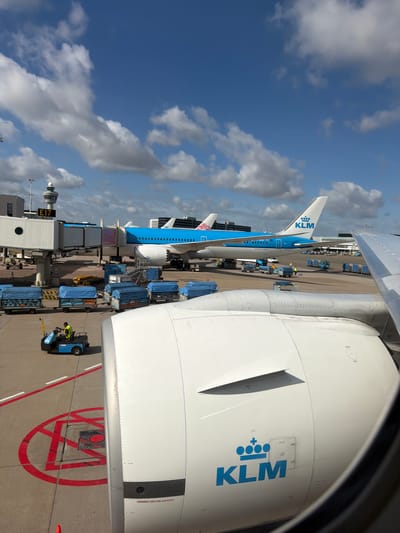 An overhead view of an airport tarmac in Badhoevedorp, Netherlands, features a KLM airplane with its distinctive blue and white livery. The plane is connected to a jet bridge at Gate E17. Several blue-covered baggage carts are lined up on the tarmac, and a small blue baggage tug with a driver wearing a high-visibility vest is in motion. In the foreground, a portion of another aircraft's engine nacelle, also in white with the KLM logo, is visible, suggesting the photo was taken from inside a plane. The sky above is a bright blue with scattered white clouds. The scene depicts typical airport operations under a clear sky.